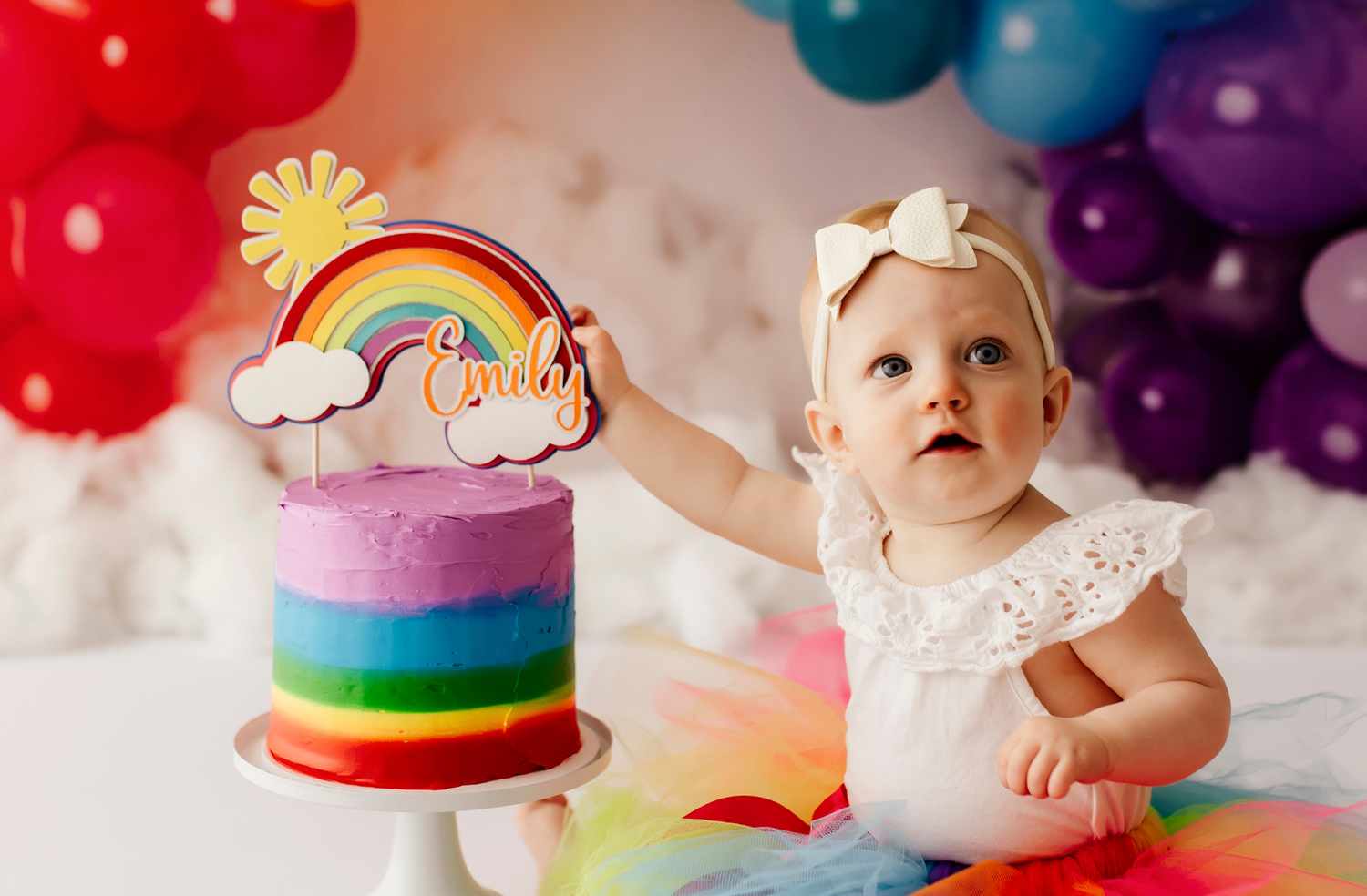 One Year Old Caucasian Baby Girl with Blue Eyes Dressed in White Frill Top and Rainbow Tutu Sitting in Front of Rainbow Balloon Backdrop Next To a Layered Rainbow Cake on a White Stand with a Rainbow Custom Cake Topper made of Layered Cardstock With a Brighth Yellow Sun Peeking out Behind the Rainbow. The name Emily is in Orange on the Cake Topper, and the Baby is Reaching for it while looking up above the camera lens. 