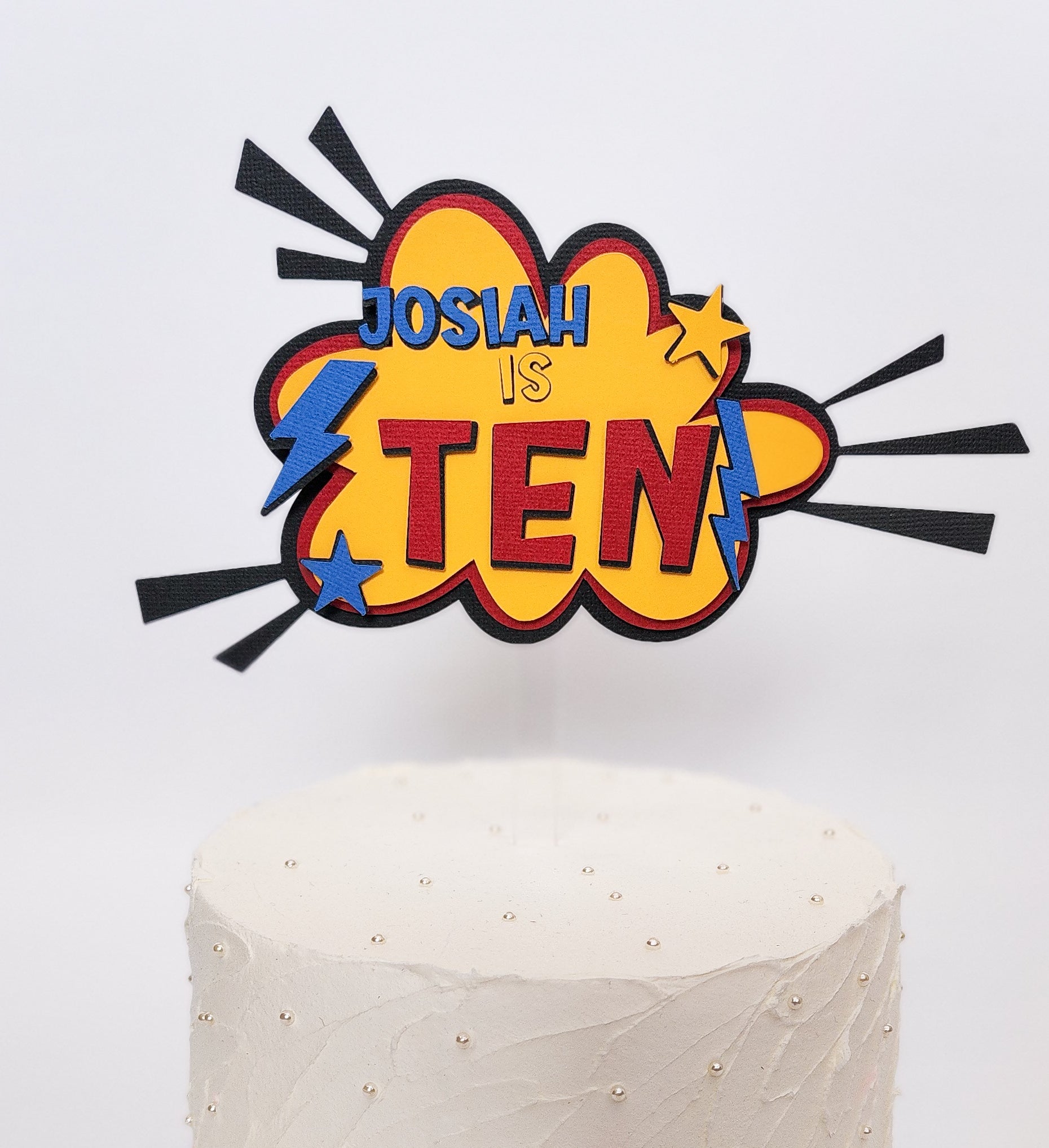 Close up photo of a superhero themed birthday cake topper made of layered cardstock. Using black, red, yellow and blue, the words "Josiah is Ten" sits on a comic style explosion background. 