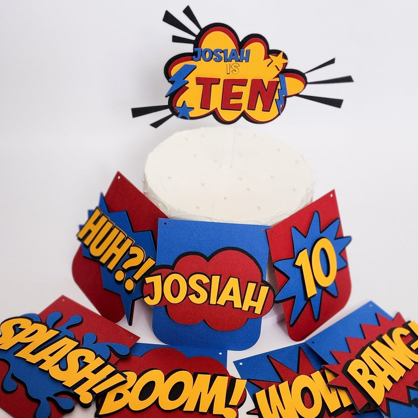 Superhero themed birthday cake topper made of layered cardstock. Using black, red, yellow and blue, the words "Josiah is Ten" sits on a comic style explosion background. 