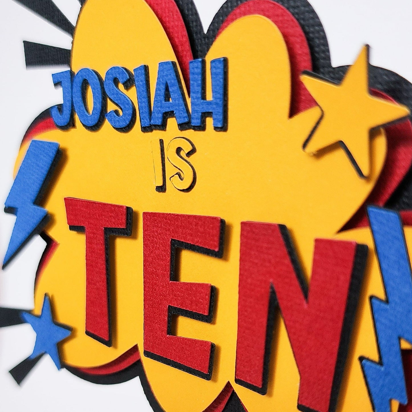 Close up photo of a superhero themed birthday cake topper made of layered cardstock. Using black, red, yellow and blue, the words "Josiah is Ten" sits on a comic style explosion background. 